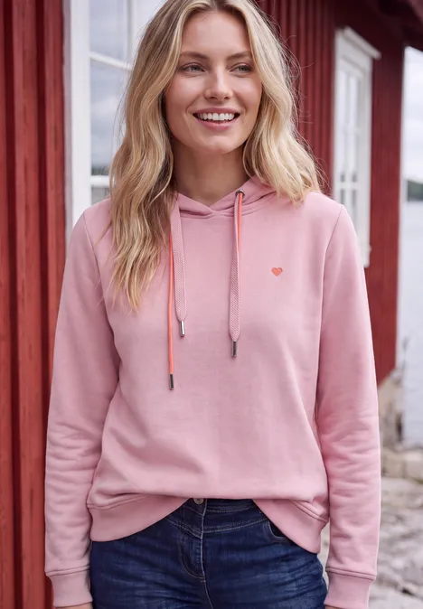Femme souriante en sweat à capuche rose et jean devant une maison rouge.