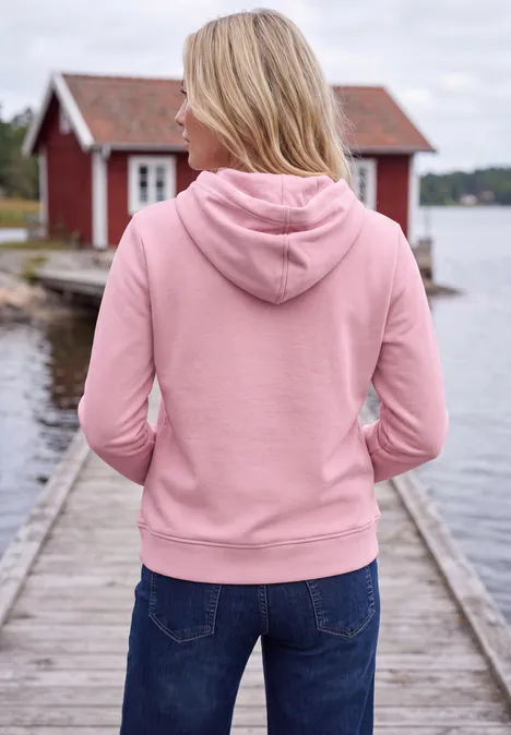 Femme en sweat à capuche rose et jean, de dos, debout sur une jetée au bord d'un lac devant une maison rouge.