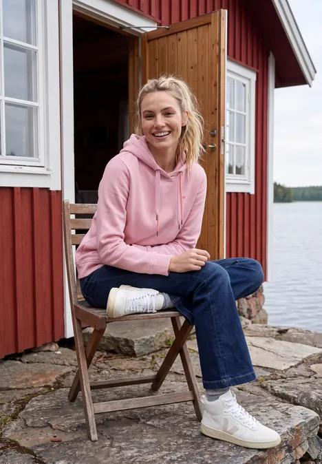 Femme en sweat à capuche rose assise sur une chaise en bois devant un chalet rouge au bord du lac.