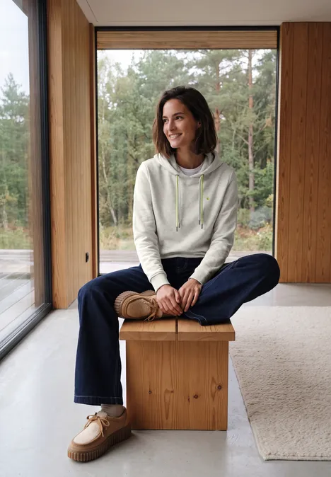 Sweat à capuche light grey melange Une femme souriante assise sur un bloc de bois dans une pièce moderne, vue sur la forêt.