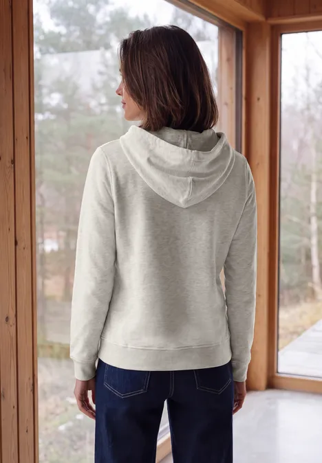 Sweat à capuche light grey melange Femme de dos en sweat à capuche gris et jean, debout devant de grandes fenêtres avec vue sur l'extérieur.