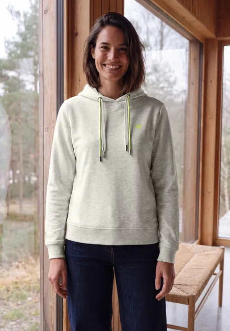 Sweat à capuche light grey melange Femme souriante en sweat à capuche gris et jean debout près d’une table en bois devant une fenêtre