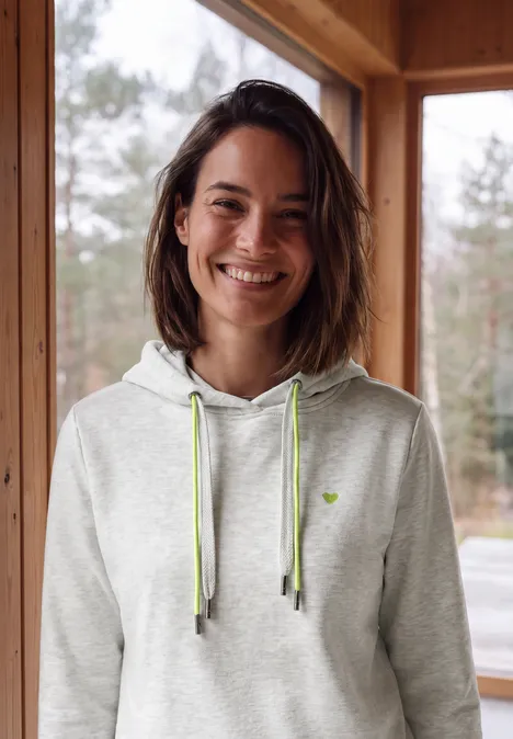 Sweat à capuche light grey melange Une femme souriante en sweat gris à capuche et cordons verts, appuyée à la fenêtre d'une cabane en bois.