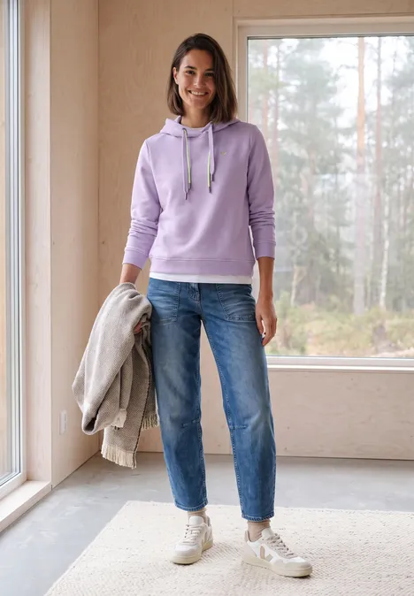 Sweat à capuche cool lilac Femme en sweat violet clair, jean et baskets, tient une veste devant une fenêtre lumineuse.