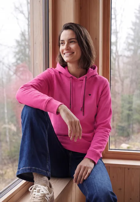 Langarm Sweatshirt mit Kapuze pink flash Frau in pinkem Kapuzenpullover und Jeans sitzt lächelnd am Fenster, Wald im Hintergrund.