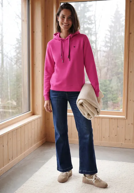 Langarm Sweatshirt mit Kapuze pink flash Frau in pinkem Hoodie, Jeans und beigen Schuhen hält Decke in einem hellen Holzraum mit großen Fenstern.