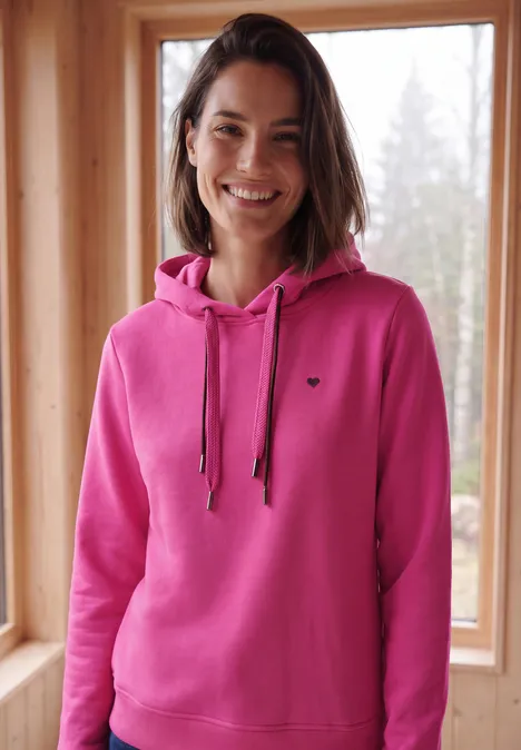 Langarm Sweatshirt mit Kapuze pink flash Frau in pinkem Kapuzenpulli, die lächelnd vor einem Fenster in einer Holzhütte steht.