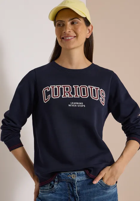 Sweatshirt mit Rundhals und Stickerei urban dark blue Frau in Jeans, dunkelblauem Sweatshirt mit Aufschrift CURIOUS und gelber Baseballmütze lächelt in die Kamera