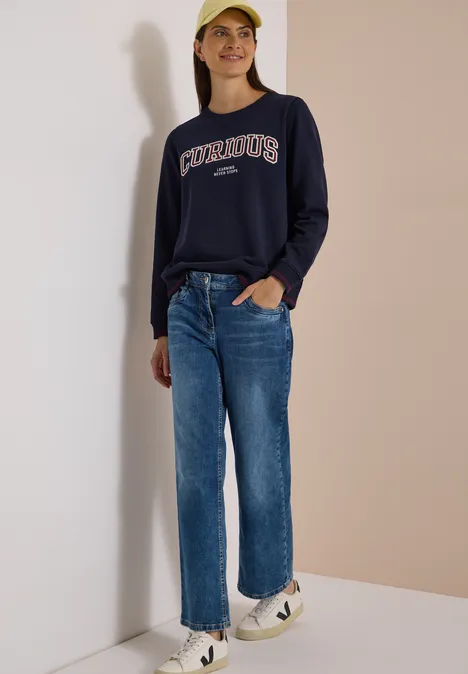 Sweatshirt mit Rundhals und Stickerei urban dark blue Junge Frau lehnt an einer Wand, trägt dunkelblauen Sweatshirt, gerade Jeans, weiße Sneakers und gelbe Mütze.