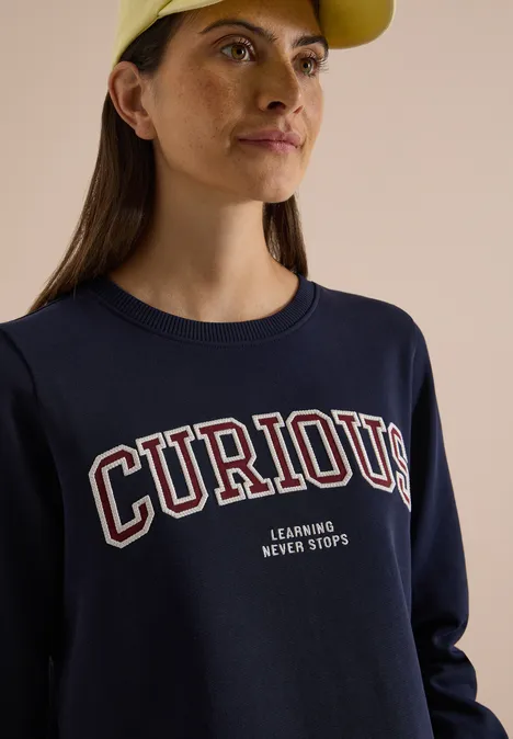 Sweatshirt mit Rundhals und Stickerei urban dark blue Eine junge Frau mit gelber Mütze und dunkelblauem Sweatshirt mit Aufschrift 'CURIOUS' blickt seitlich.