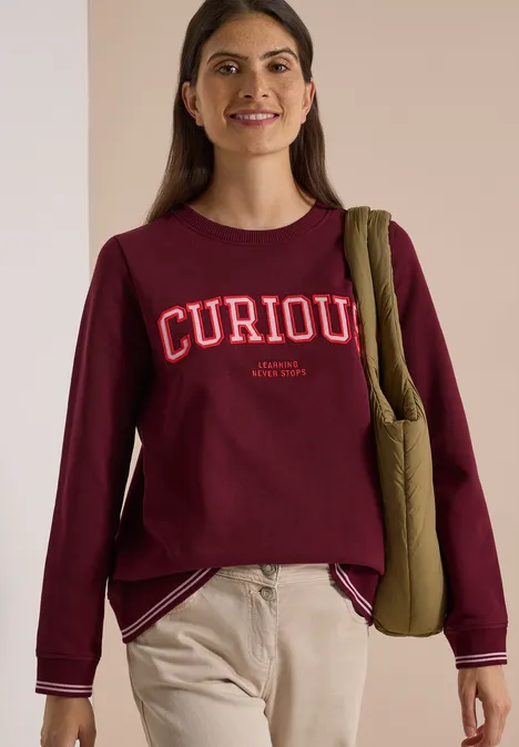 Frau trägt burgunderfarbenes Sweatshirt mit Aufschrift 'CURIOUS', beige Hose und grünen Schulterbeutel.