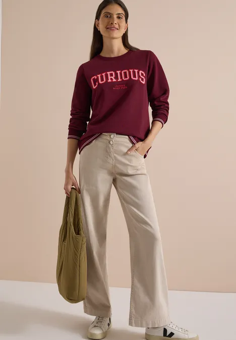 Frau trägt bordeauxfarbenes Sweatshirt mit 'CURIOUS', helle weite Hose, olivfarbener Beutel, weiße Sneaker