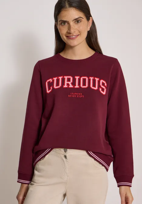 Frau mit weinrotem Sweatshirt mit 'CURIOUS'-Aufschrift und beigen Hosen, lächelt in die Kamera.