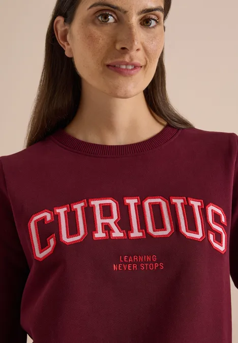 Frau in bordeauxrotem Pullover mit großem „CURIOUS“-Schriftzug und kleinem „Learning Never Stops“-Motto lächelnd