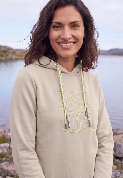 Eine lächelnde Frau im beigen Hoodie steht vor einem See.