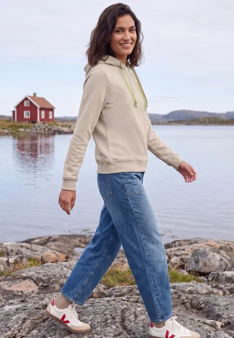 Eine Frau in Hoodie und Jeans spaziert am Seeufer vor einer roten Hütte.