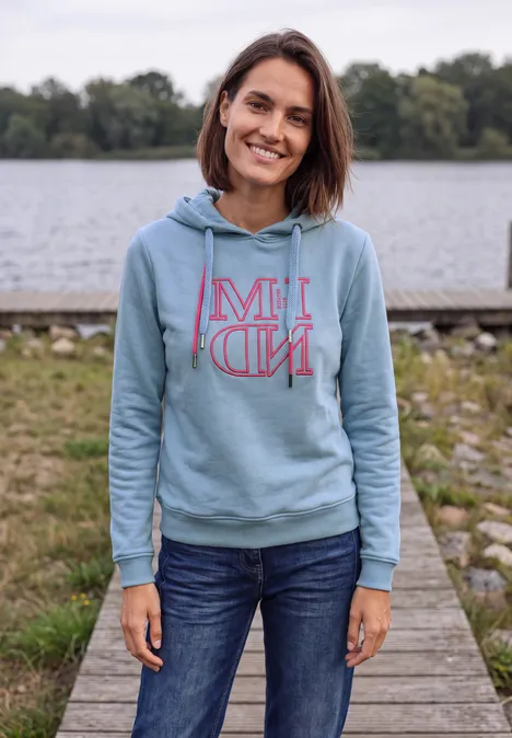 Eine lächelnde Frau in Jeans und hellblauem Hoodie steht auf einem Steg an einem See.