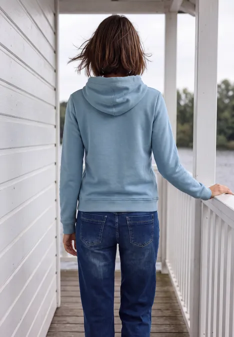 Person in hellblauer Kapuzenjacke und Jeans steht auf einem weißen Balkon, blickt in die Natur.