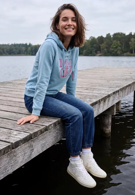 Frau lächelt auf einem Steg am See, trägt Hoodie, Jeans und weiße Schuhe.