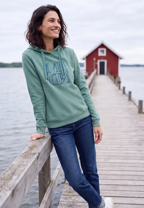 Sweatshirt met capuchon en Artwork mineral blue Eine Frau lehnt lächelnd an einem Steg am See, trägt grünen Hoodie und Jeans, im Hintergrund eine rote Bootshütte.
