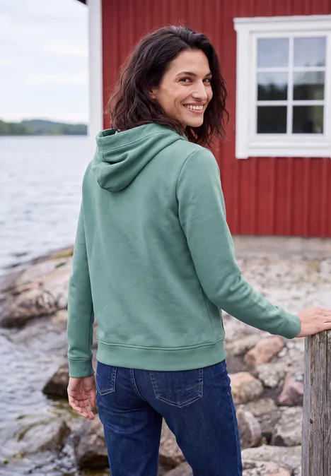 Sweatshirt met capuchon en Artwork mineral blue Eine lächelnde Frau in grüner Kapuzenjacke und Jeans steht am Ufer vor einem roten Haus am See.