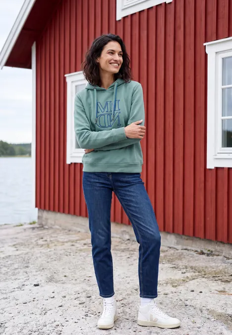 Sweatshirt met capuchon en Artwork mineral blue Frau in grüner Kapuzenjacke, Jeans und weißen Stiefeletten steht vor roter Holzhütte am See.