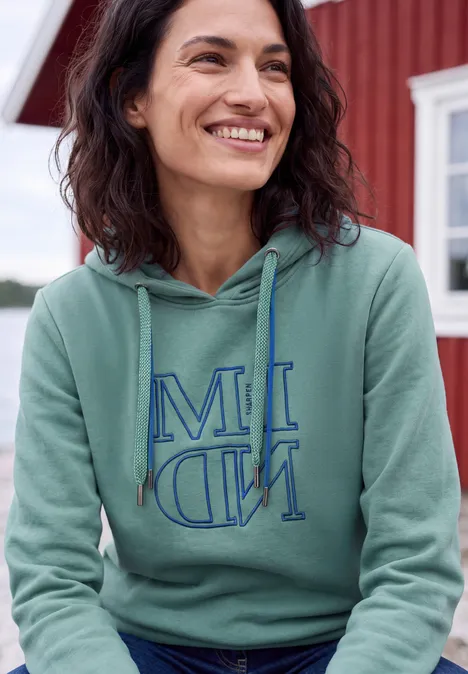 Sweatshirt met capuchon en Artwork mineral blue Eine lächelnde Frau in grünem Kapuzenpullover vor einem roten Haus