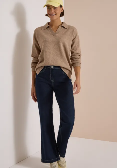 Frau in beige Pullover, weiten Jeans, weißen Sneakern und gelber Cap vor neutralem Hintergrund.