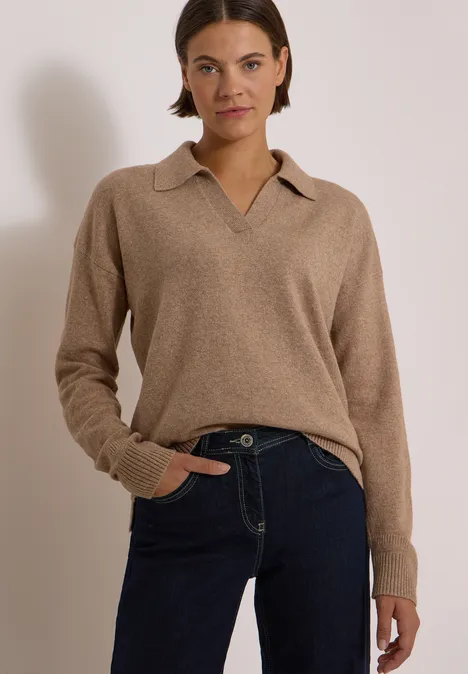 Frau trägt beigen Polopullover mit Kragen und dunkle Jeans vor hellem Hintergrund.