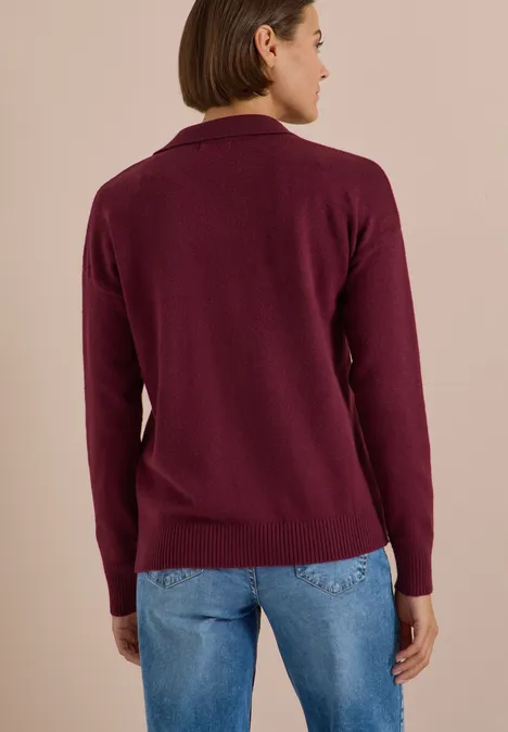 Rückansicht einer Person in dunkelrotem Strickpullover und blauen Jeans vor einfarbigem Hintergrund.