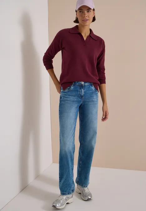 Frau trägt dunkelrotes Poloshirt, blaue Jeans, weiße Sneakers und rosa Cap vor neutralem Hintergrund.