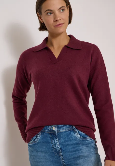 Frau in burgunderfarbenem Pullover mit V-Kragen und blauen Jeans posiert vor weißem Hintergrund.