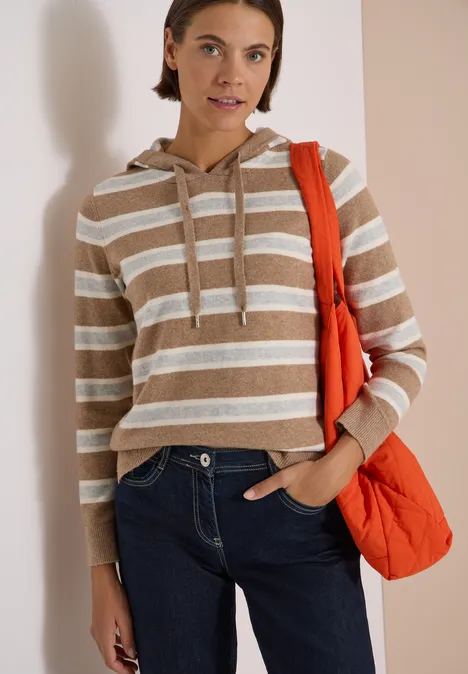 Junge Frau trägt gestreiften Kapuzenpullover, Jeans und orangefarbene Tasche vor hellem Hintergrund.