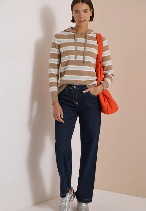 Frau in gestreiftem Kapuzenpullover, blauen Jeans und roter Umhängetasche vor hellem Hintergrund.