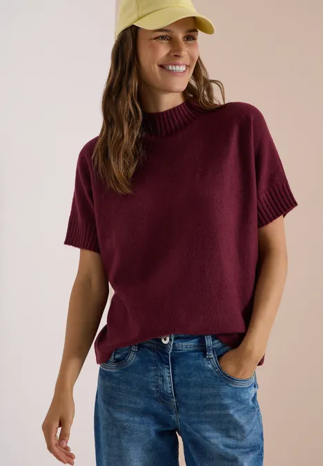 Dolman-trui met col red wine Frau in lässigem Kurzarm-Pullover mit hohem Kragen in Bordeaux, Jeans und gelber Kappe lächelnd.