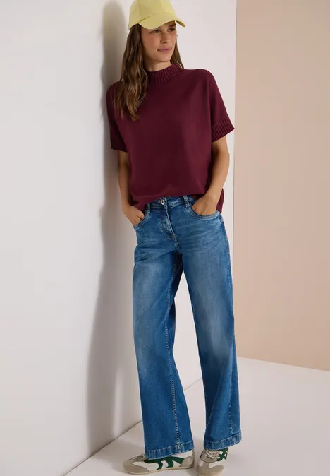 Dolman-trui met col red wine Frau lehnt an einer Wand, trägt ein bordeauxfarbenes T-Shirt, weite Jeans und eine gelbe Kappe.