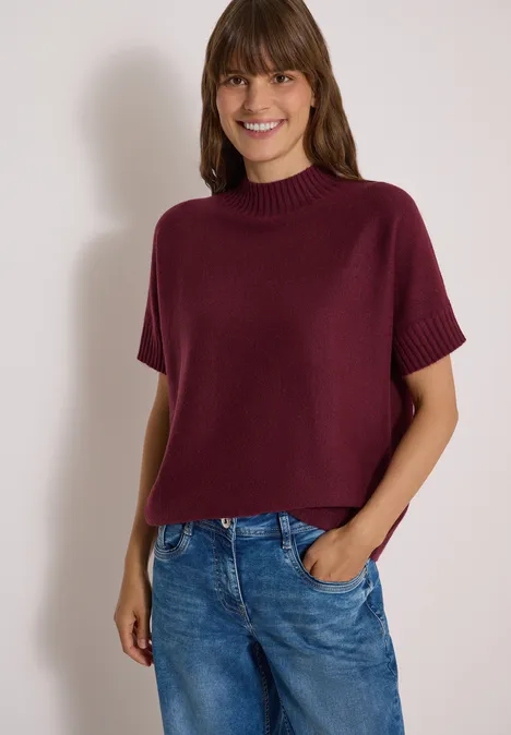 Dolman-trui met col red wine Frau in kurzärmligem, bordeauxrotem Strickpullover und Jeans lächelt vor hellem Hintergrund.