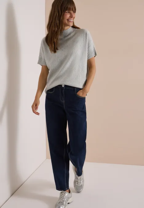 Dolman-trui met col light grey melange Frau lehnt lässig an Wand und trägt graues T-Shirt, blaue Jeans und weiße Sneaker.