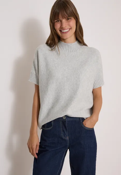 Dolman-trui met col light grey melange Junge Frau mit braunem Haar, grauem Kurzarm-Pullover und Jeans, lächelnd vor hellem Hintergrund.