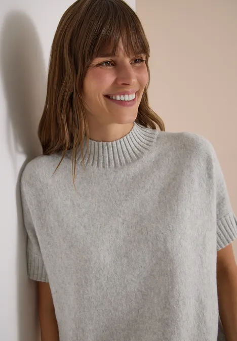 Dolman-trui met col light grey melange Eine lächelnde Frau lehnt an einer Wand und trägt einen grauen Kurzarmpullover.