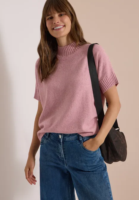 Dolman-trui met col velvet rose melange Frau in rosa Kurzarm-Pullover, Jeans und Schultertasche steht lächelnd vor neutralem Hintergrund.