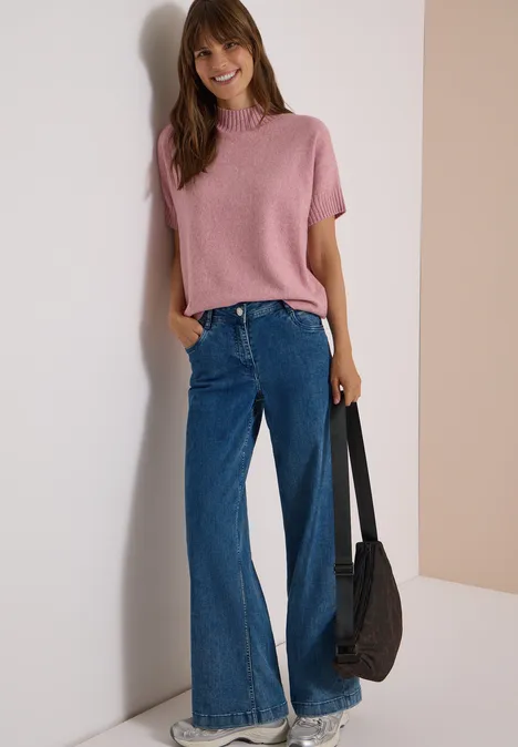 Dolman-trui met col velvet rose melange Frau lehnt lächelnd an Wand, trägt rosa Kurzarm-Pullover, weite Jeans und hält schwarze Tasche.