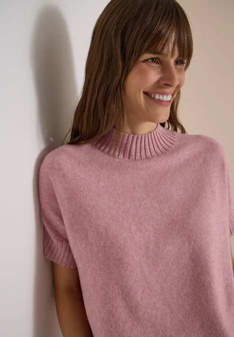 Dolman-trui met col velvet rose melange Eine lächelnde Frau lehnt an einer Wand und trägt einen rosa Kurzarm-Pullover.