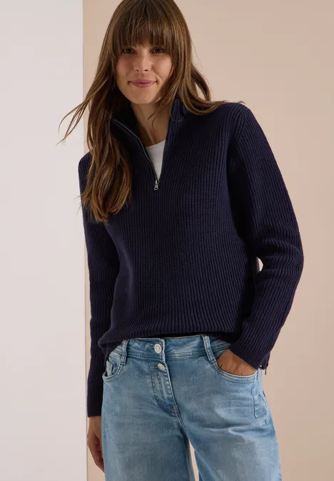 Frau mit halblangem dunkelblauem Pullover mit Reißverschluss und Jeans, posierend vor neutralem Hintergrund.