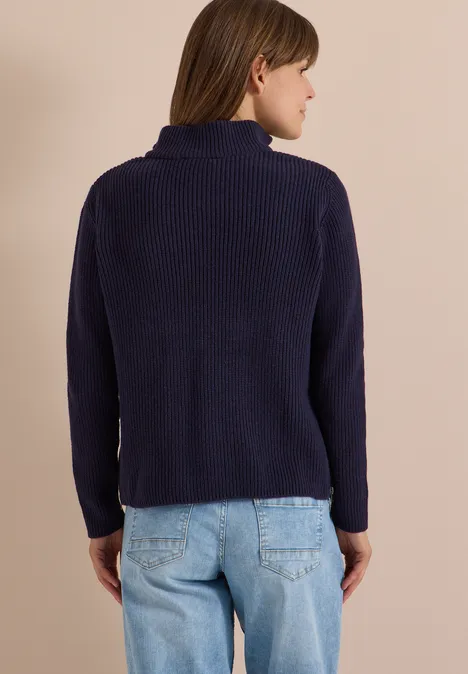 Frau von hinten in dunkelblauem Strickpullover mit hohem Kragen und hellen Jeans.