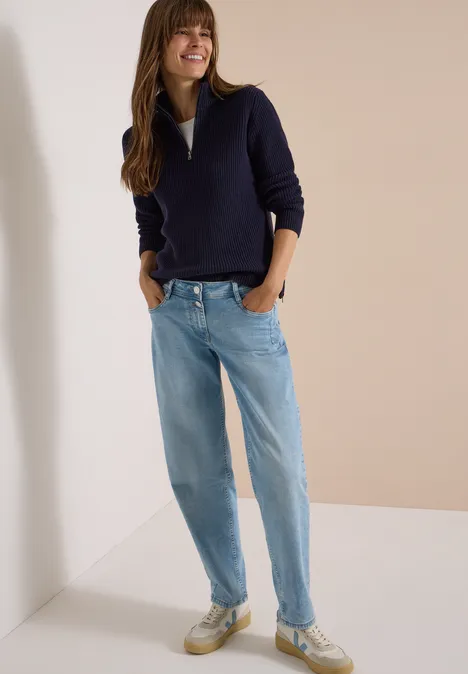 Frau trägt dunkelblauen Pullover, helle Jeans und Sneaker, steht lächelnd.