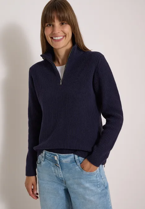 Lächelnde Frau in navyblauem Pullover und Jeans, eine Hand in der Hosentasche.
