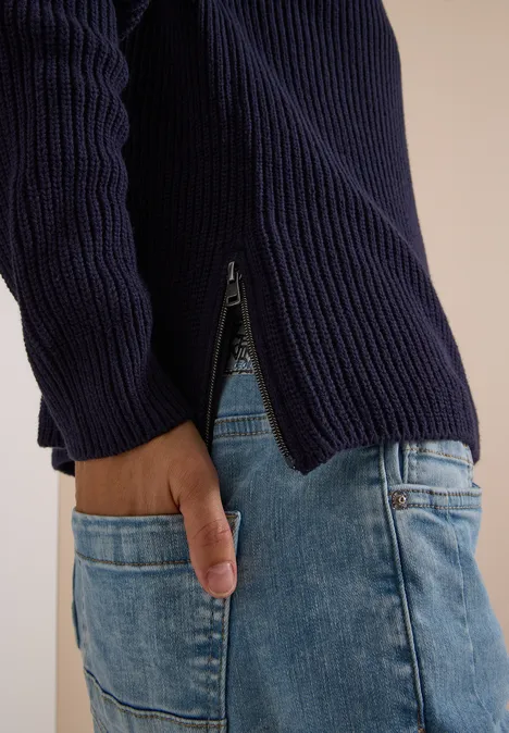 Nahaufnahme eines Oberkörpers in dunkelblauem Rippsweater mit seitlichem Reißverschluss und hellen Jeans.