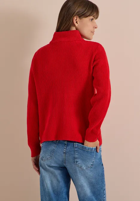 Frau von hinten, schaut seitlich nach rechts, trägt roten Rollkragenpullover und blaue Jeans vor neutralem Hintergrund.