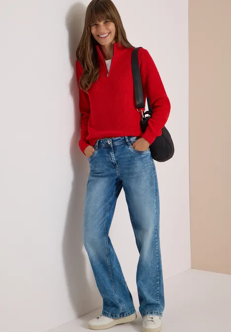 Eine Frau in rotem Pullover, blauen Jeans und Sneakern lehnt lächelnd an einer Wand, trägt eine Tasche.